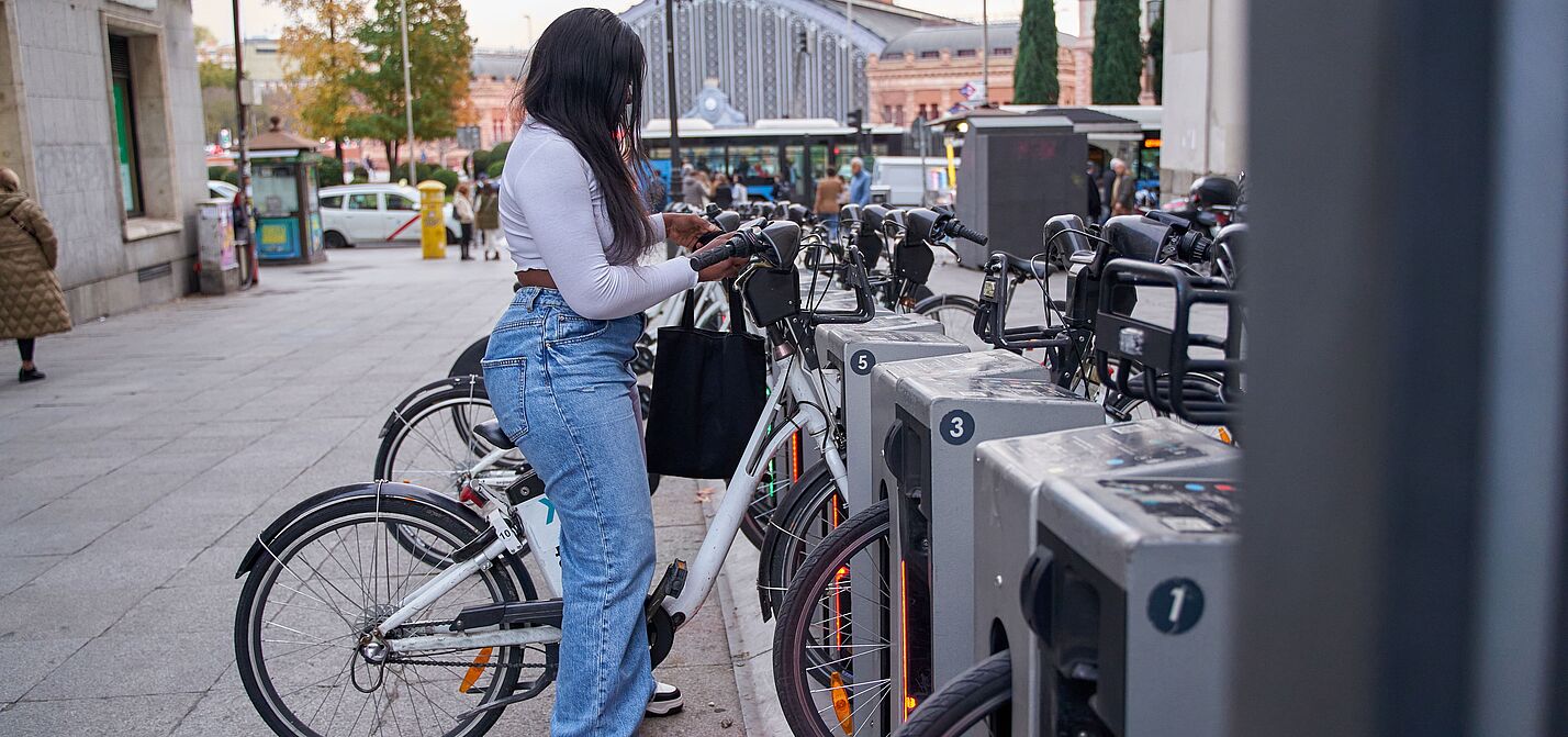 Eine junge Frau mit langen dunklen Haaren, weißem Oberteil, Jeans und schwarzer Umhängetasche steht an einer Bike-Sharing-Station und entnimmt ein weißes Leihfahrrad. Die Station verfügt über mehrere nummerierte Stellplätze mit grauen Dockingelementen. Im Hintergrund sind ein belebter Stadtplatz, historische Gebäude, Passanten und ein Bahnhofsgebäude zu sehen.