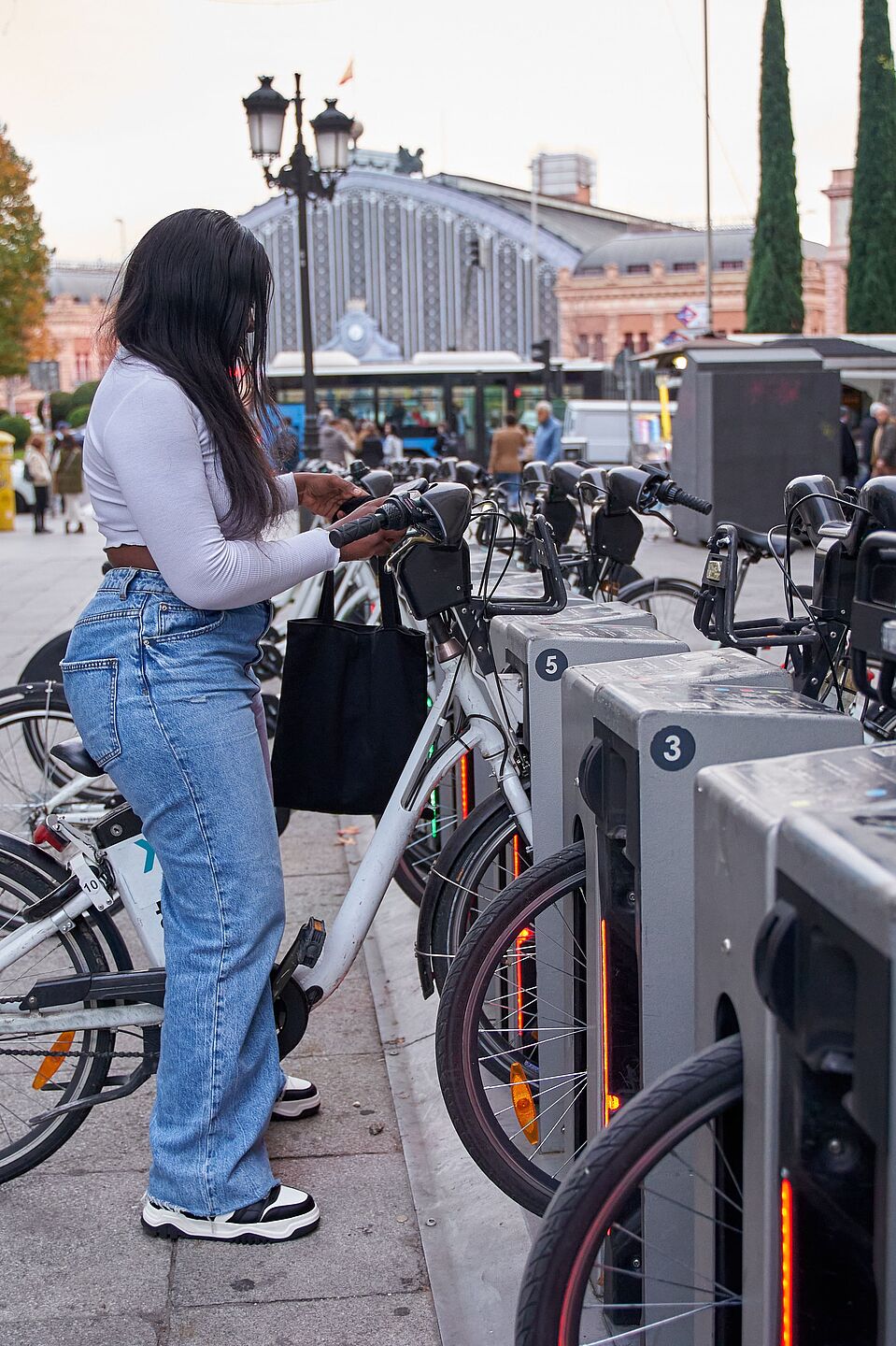 Frau entleiht Fahrrad an Bikesharing-Station Eine junge Frau mit langen dunklen Haaren, weißem Oberteil, Jeans und schwarzer Umhängetasche steht an einer Bike-Sharing-Station und entnimmt ein weißes Leihfahrrad. Die Station verfügt über mehrere nummerierte Stellplätze mit grauen Dockingelementen. Im Hintergrund sind ein belebter Stadtplatz, historische Gebäude, Passanten und ein Bahnhofsgebäude zu sehen.